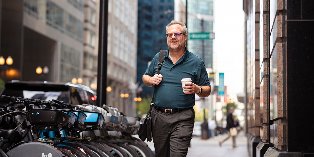 Man carrying a messenger bag and a cup of coffee walking down a busy street.