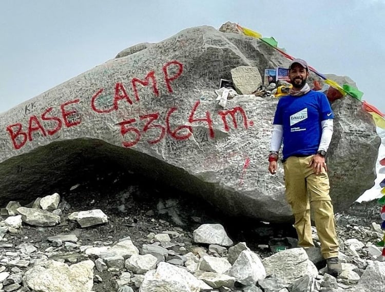 Karan Saberwal stands in his Baird shirt next to a large rock with "Base Camp 5364m." spray painted on it