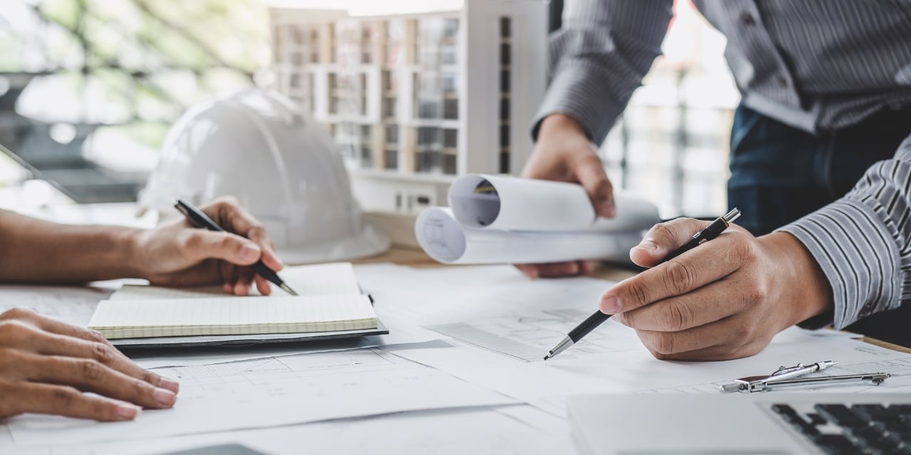 Two people reviewing architectural plans on a desk with hard hat and building model in background.