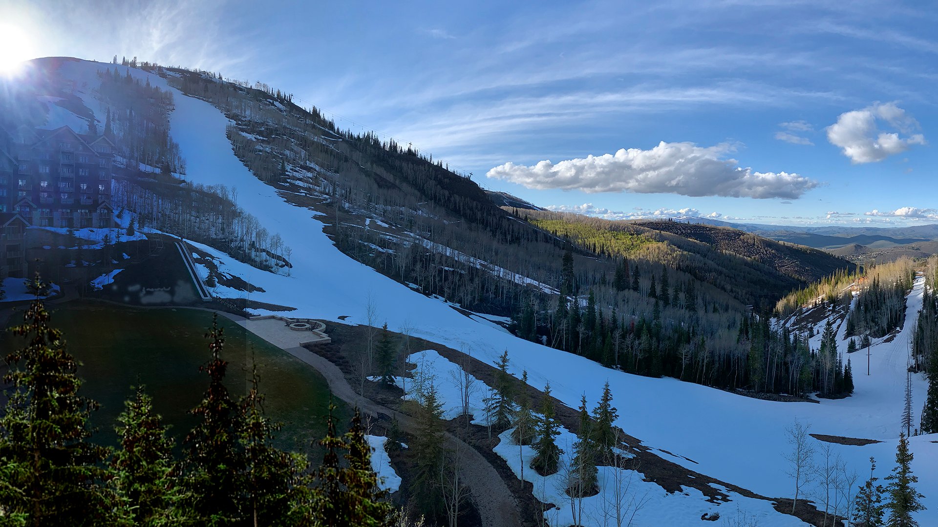 Snow‑covered mountain slopes with scattered trees under a bright sky, showing a resort area transitioning from winter.