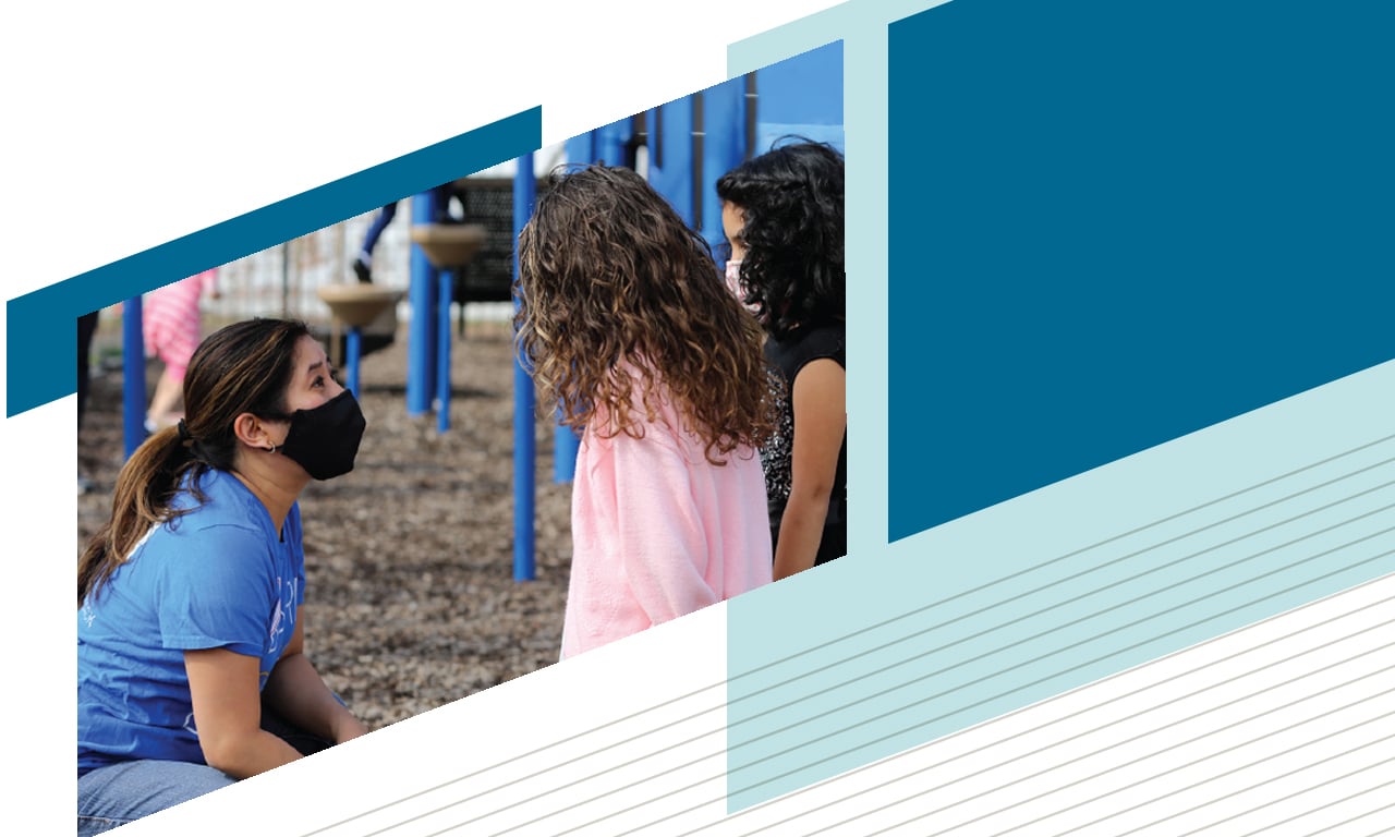 An image of a woman in a Baird Gives Back t-shirt talking to two school children on a playground while she volunteers