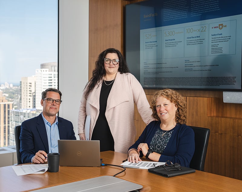Three associates from Baird Equity Capital Markets around a table in a conference room with a presentation on screen in the background