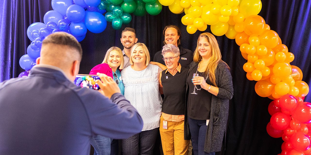 A group of Baird associates pose for a photo under an arch of rainbow colored balloons at a corporate event