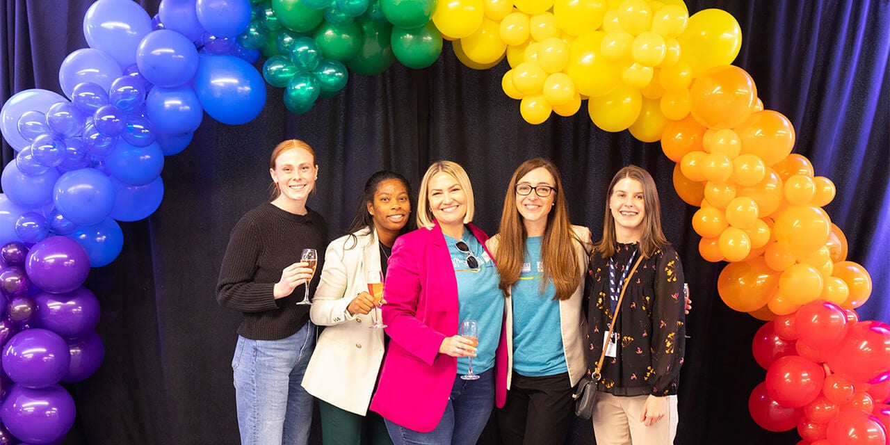 A group of Baird associates stand below an arch of rainbow balloons at a Spectrum ARG event