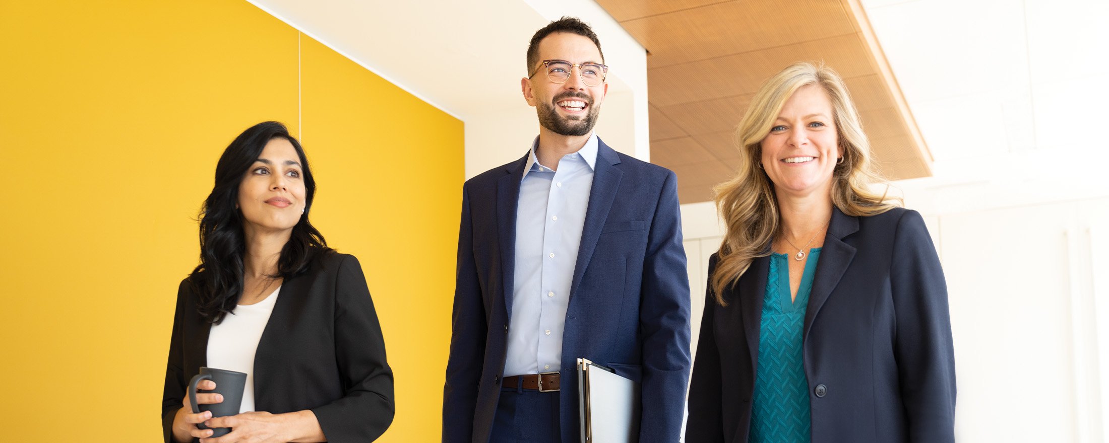 Three people stand in a brightly lit hallway with a yellow wall on the left