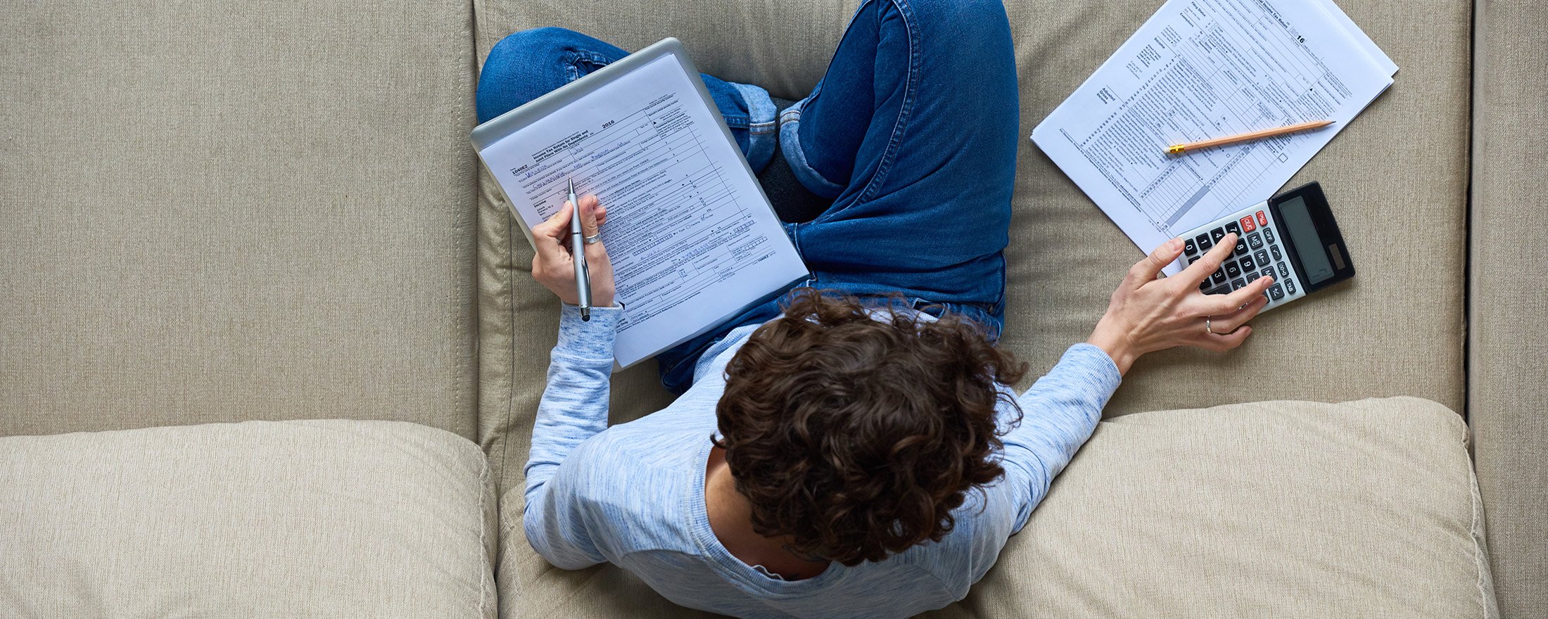 Overhead view of person reviewing financial documents with calculator and pen on a sofa