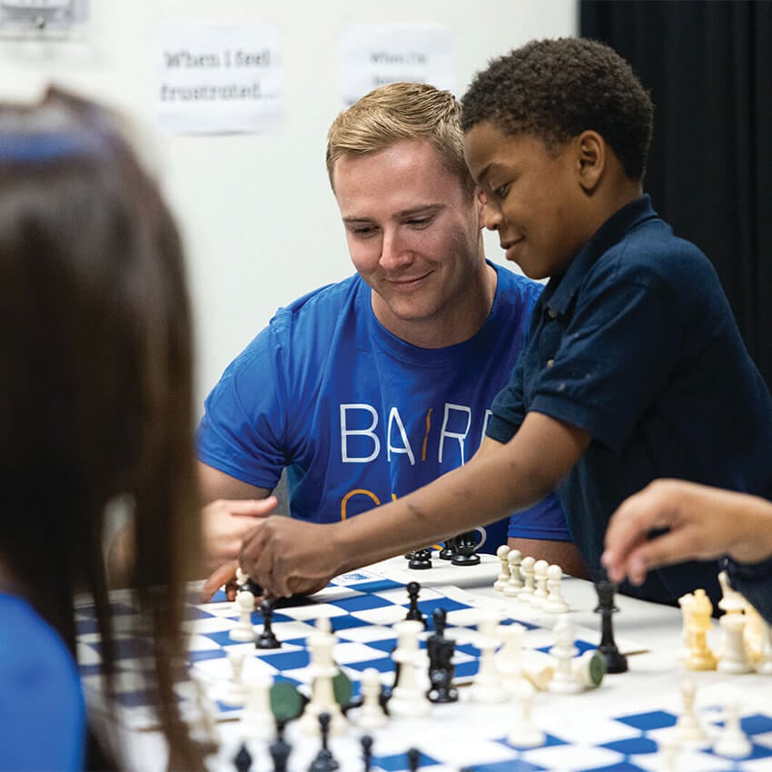 A man in a Baird Gives Back t-shirt is helping a child learn how to play chess.