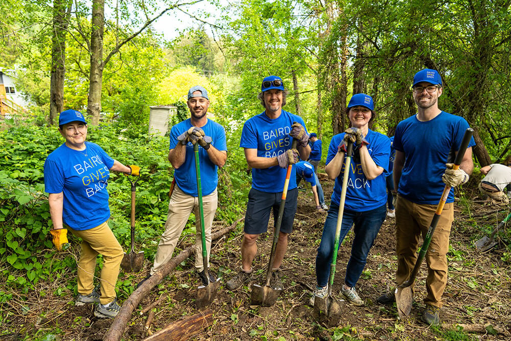 Five Baird associates wearing Baird Gives Back t-shirts and hats and holding shovels at Longfellow Creek in Seattle