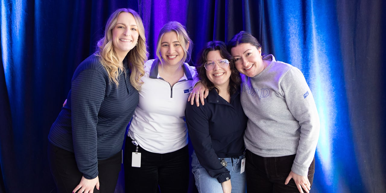 Four women standing in front of a blue and purple backdrop smiling. 
