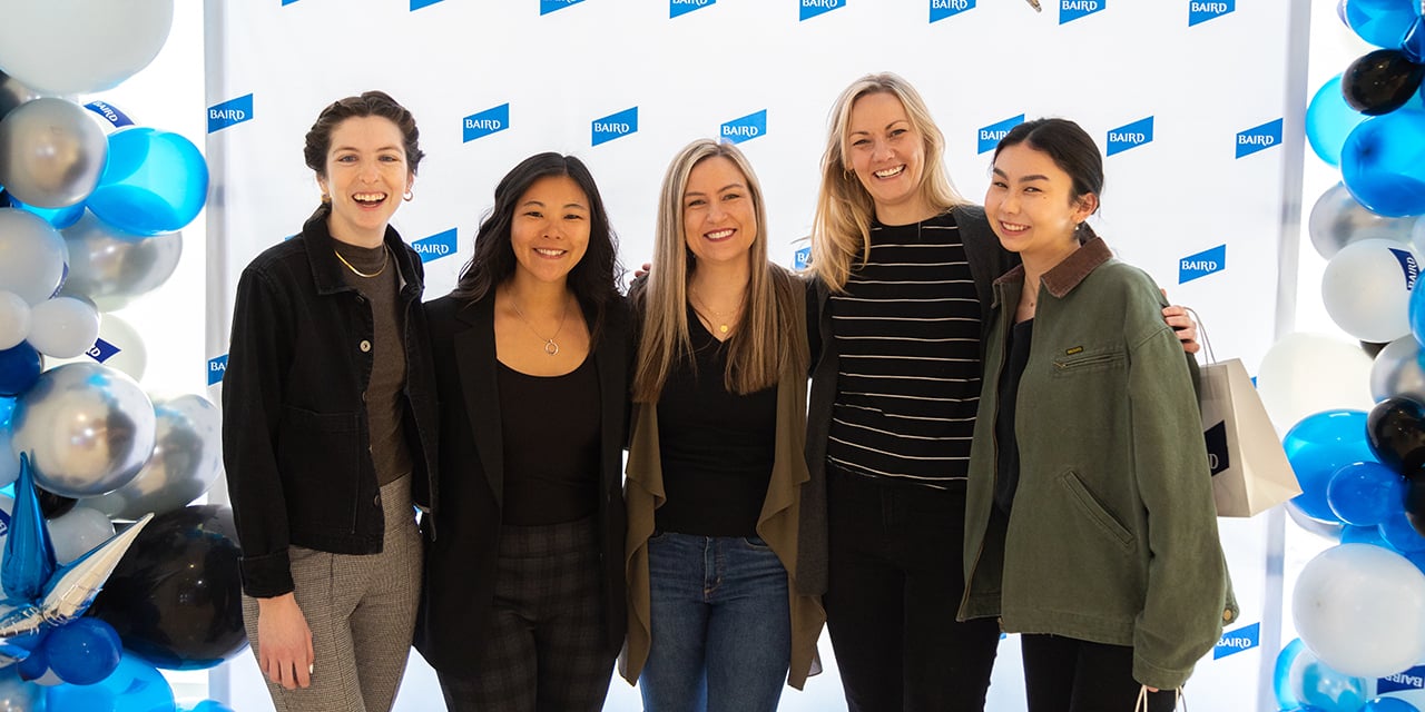 Five women stand before a Baird backdrop with blue, white, and silver balloons.