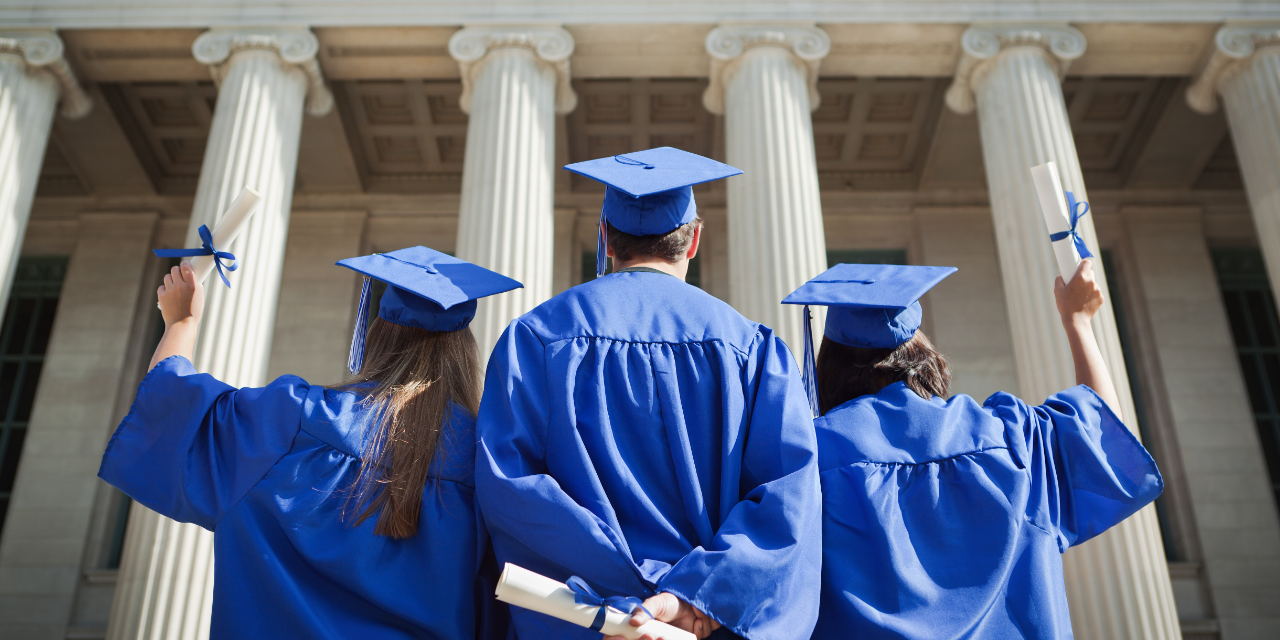 Students in blue graduations gowns and caps standing in front of a historic educational building.