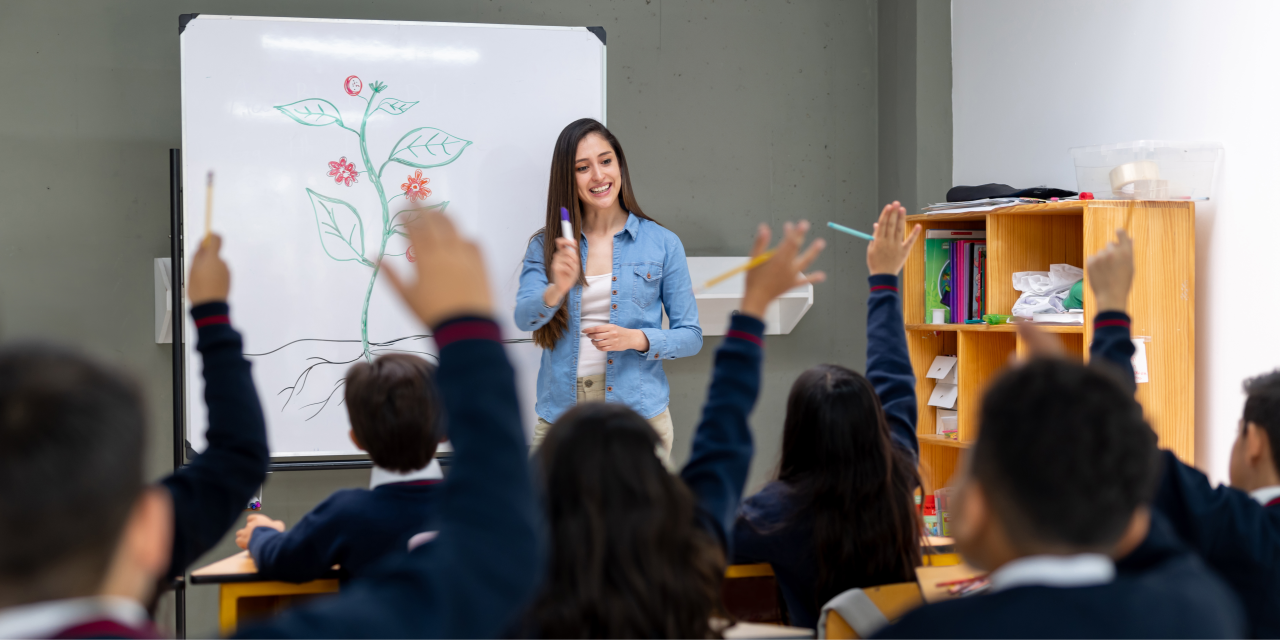 Teacher at whiteboard in front of a classroom with children wearing uniforms and raising hands.