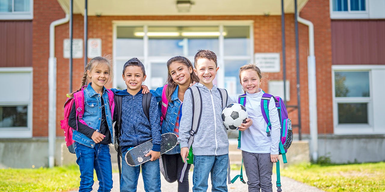 Five children with backpacks standing together outside a school entrance, holding a soccer ball and skateboard.