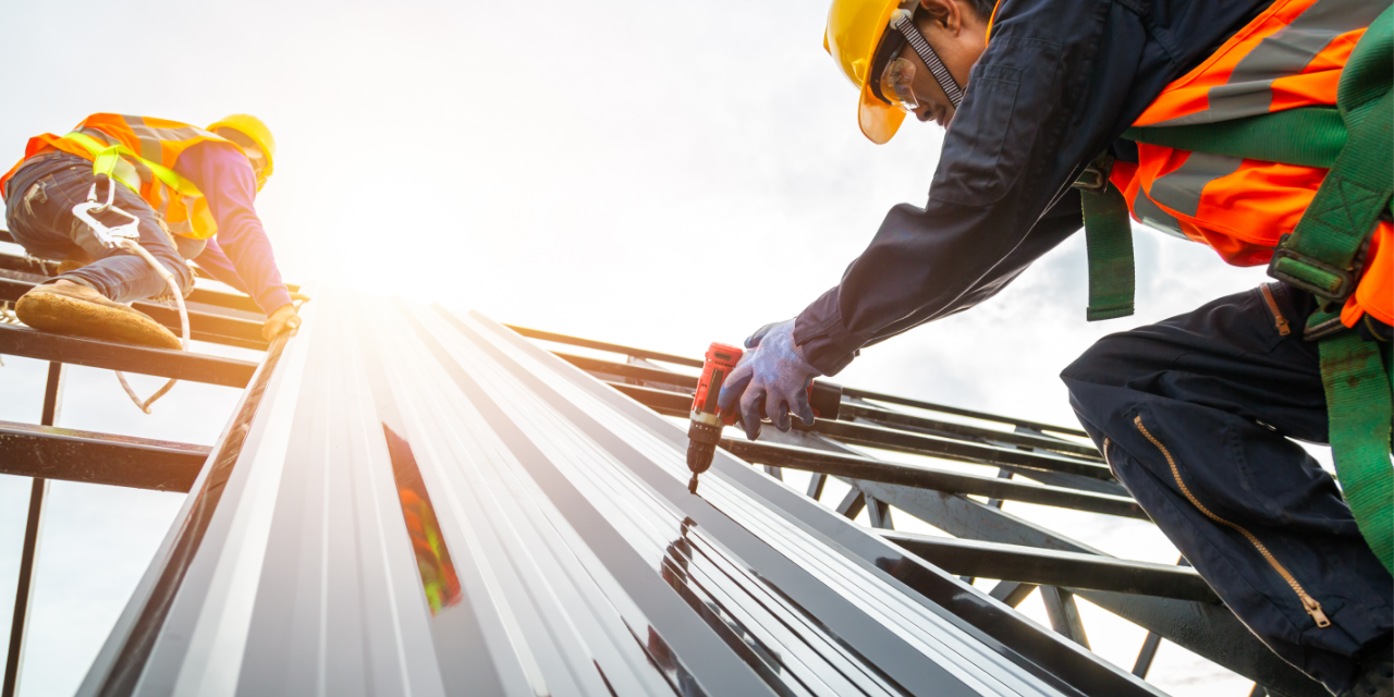 Workers install metal roofing panels using power tools on a construction site under bright sunlight.
