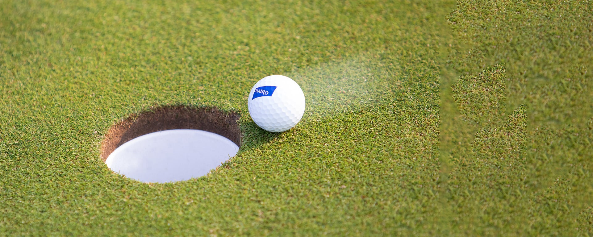 A golf ball printed with the Baird logo moving towards a hole on the green