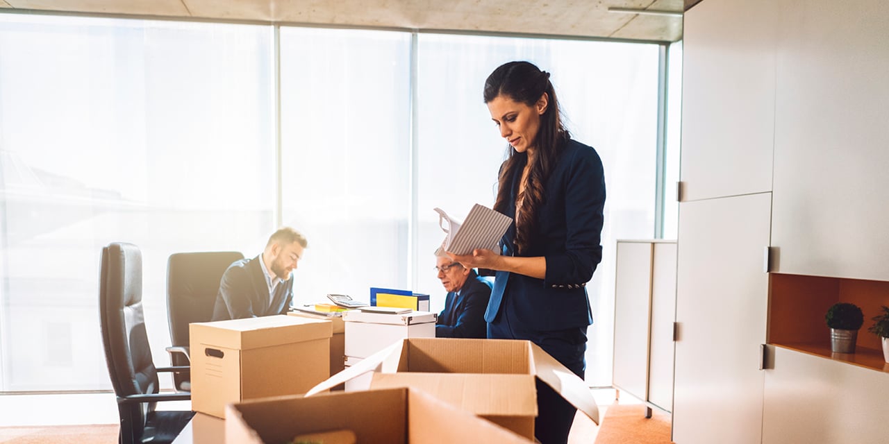 Three professionals in a bright office; one organizes boxes, others work at desks with computers.