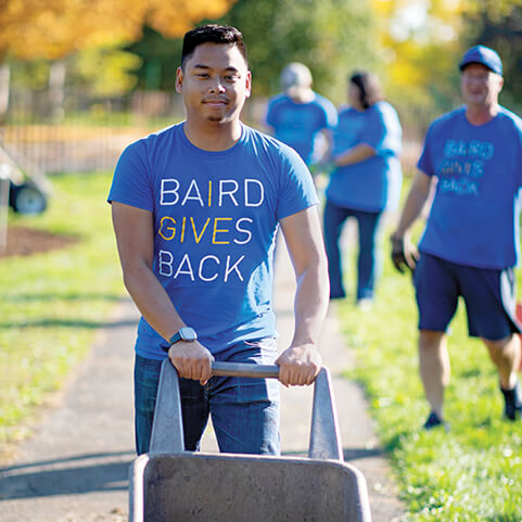 A candid photo of a man pushing a wheelbarrow and wearing a Baird Gives Back t-shirt with other Baird volunteers out of focus in the background