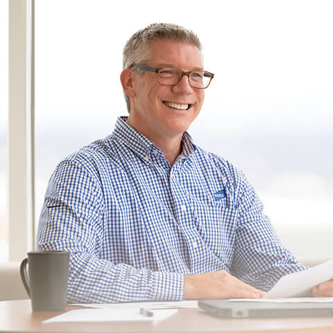 A smiling man sits at a desk with a laptop, cup of coffee, and documents spread out in front of him