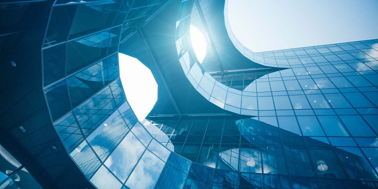 Curved glass building facade reflecting blue sky and sunlight through an open circular structure.