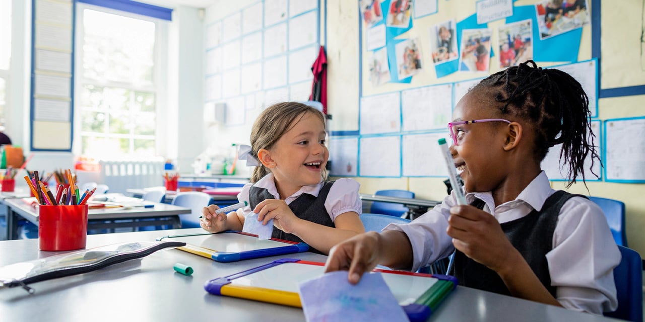 Two elementary school children wearing uniforms and laughing while working at desks.