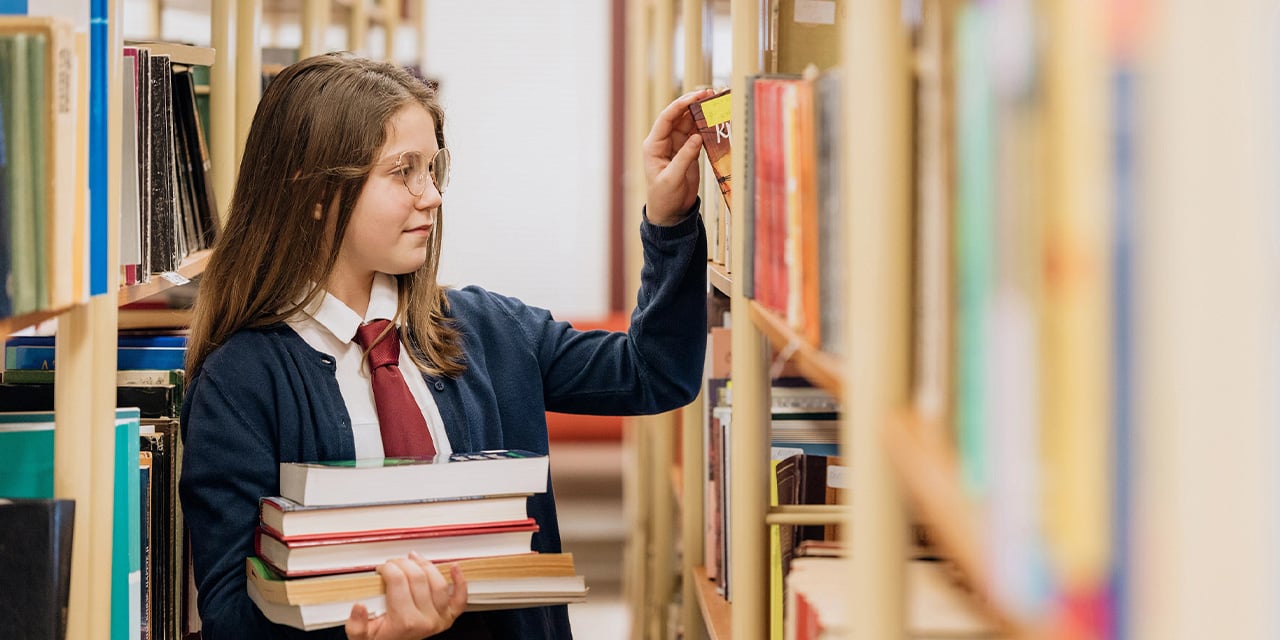 Student carrying a stack of books while looking at additional books on shelves in a library.