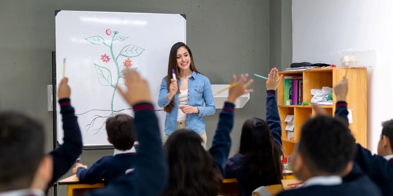 View form the back of a classroom where students in uniforms are raising their hands.