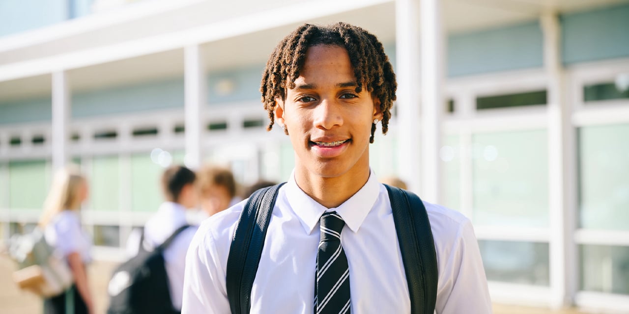 Male high school student wearing a uniform and carrying a backpack standing outside of a school building.