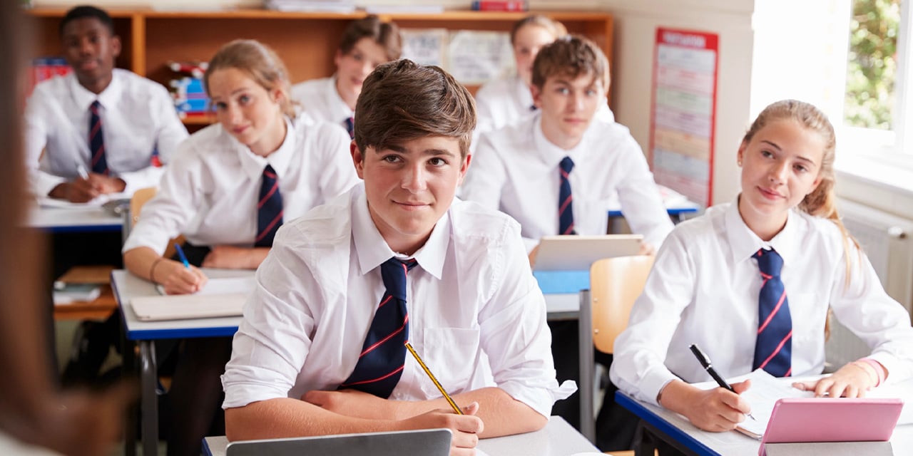 High school students earing white shirt and tie uniforms sitting at desks in a classroom.