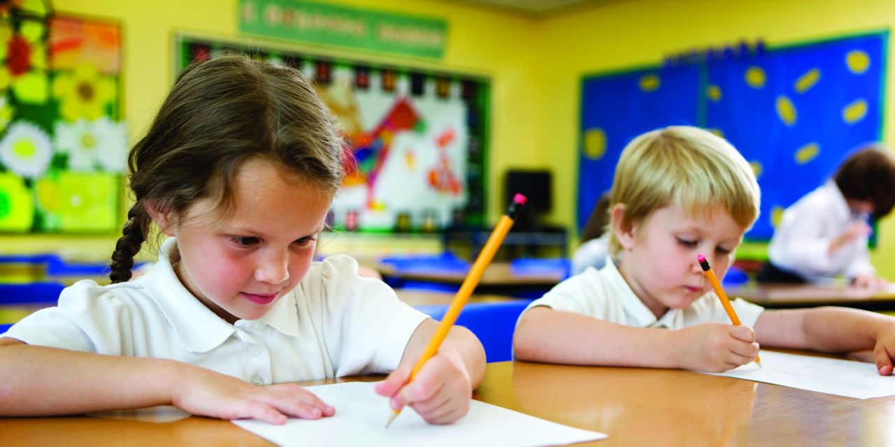 Two elementary school students sitting at shared desk while writing on paper with a pencil.