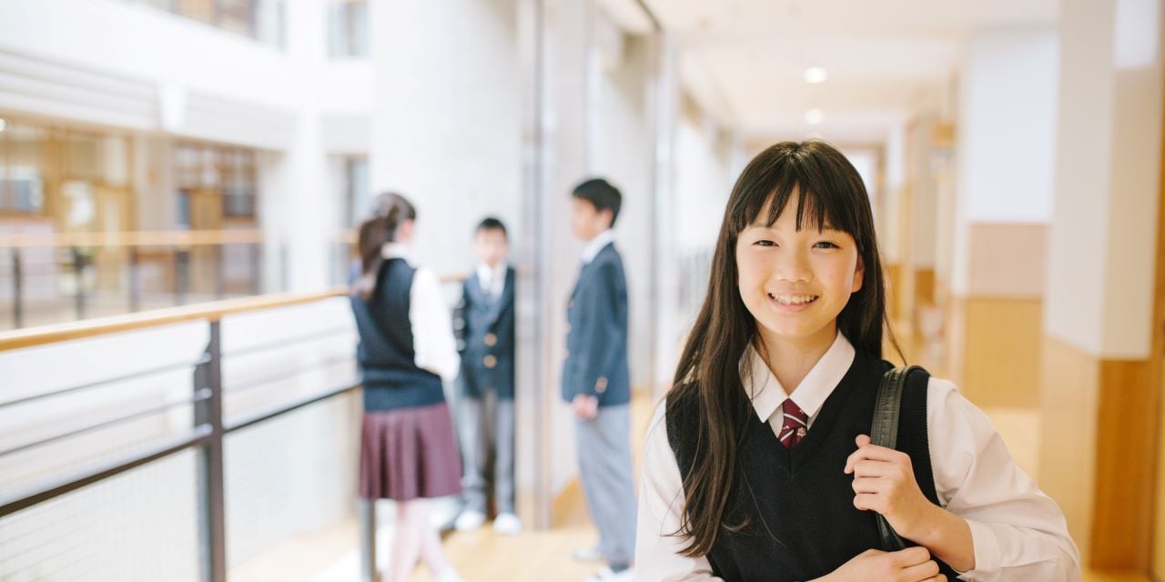 Student smiling and holding a backpack while other students congregate in the hallway in the background.