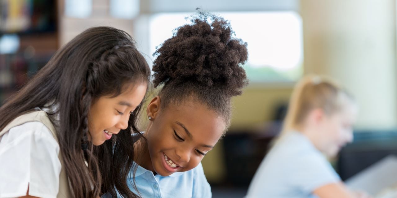 Two students sitting together looking at something on a desk with smiles on their faces.