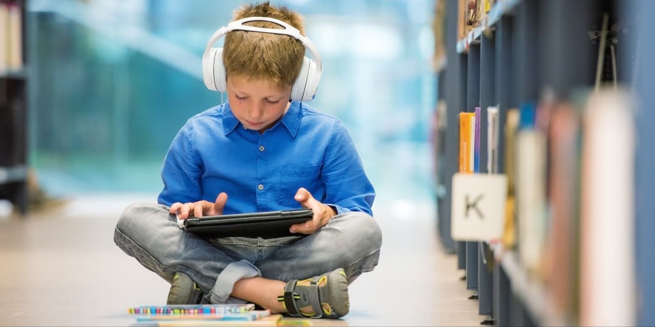 Elementary school aged child sitting in a library with headphones on head and looking at an ipad.