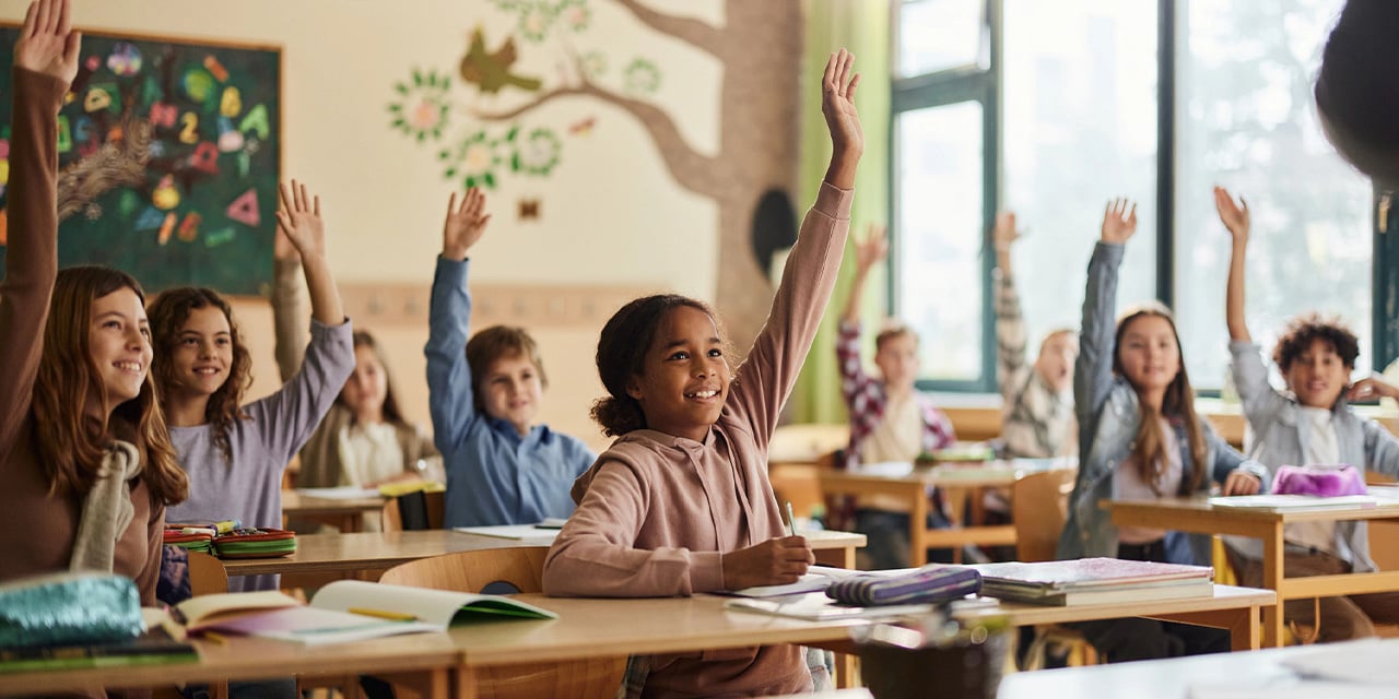Students in a classroom raising their hands while sitting at desks.