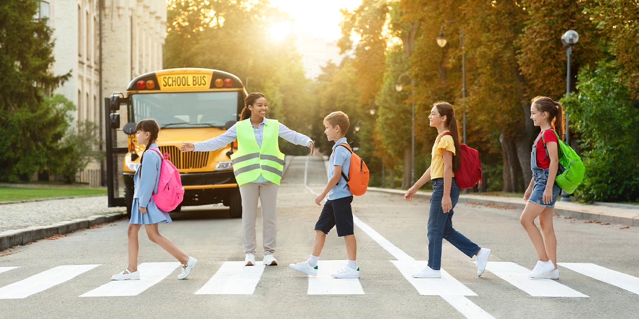 Students being assisted by a crossing guard as they cross a street as a bus waits in the background.