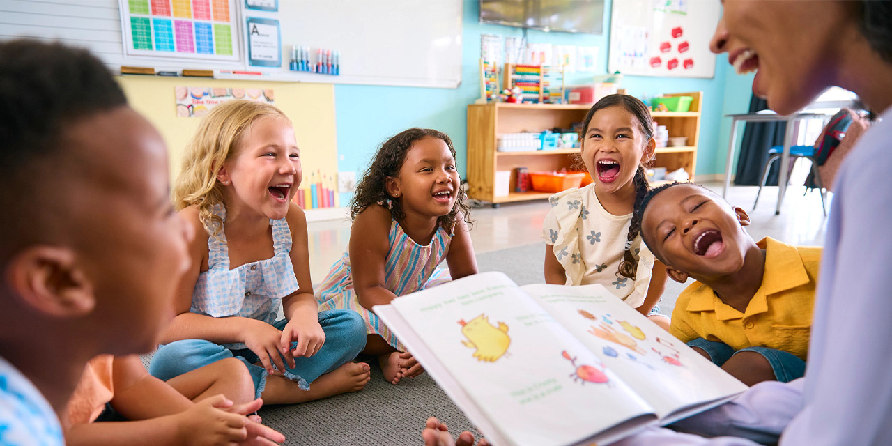 Elementary school ages children sitting on a carpet laughing while listening to an adult read them a story.