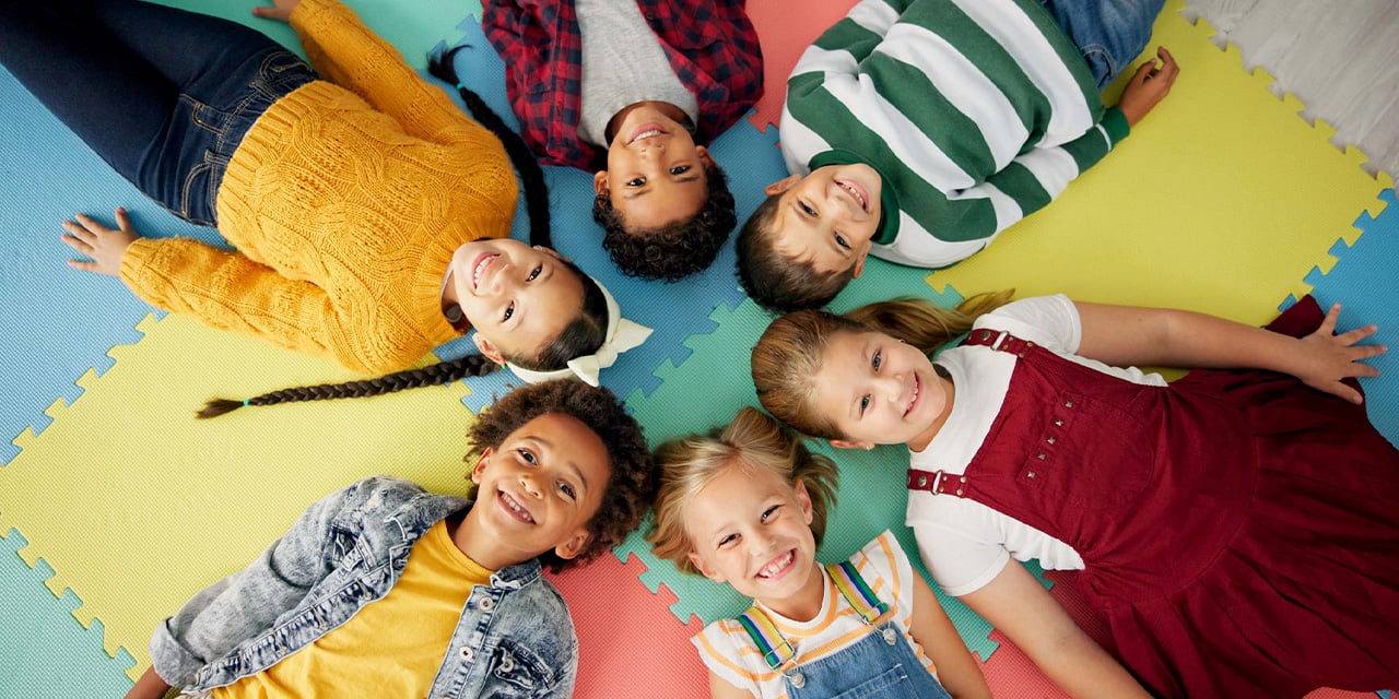 Elementary school ages children laying on a carpet with their heads together in a circle.