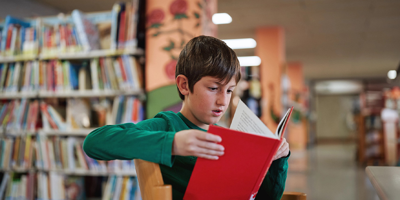 Elementary school student reading a book while sitting in a library.