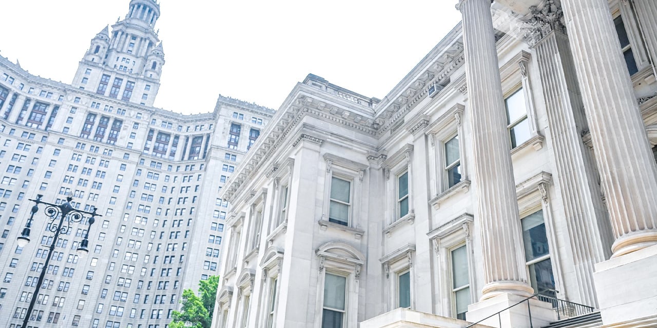 Historic stone building with tall columns beside a large ornate high-rise against a bright sky.