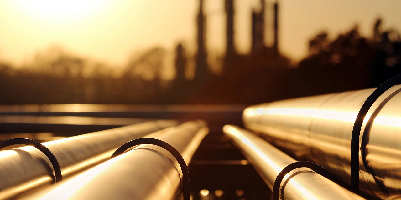 Metal pipelines glowing in warm sunset light with an industrial facility blurred in the background.