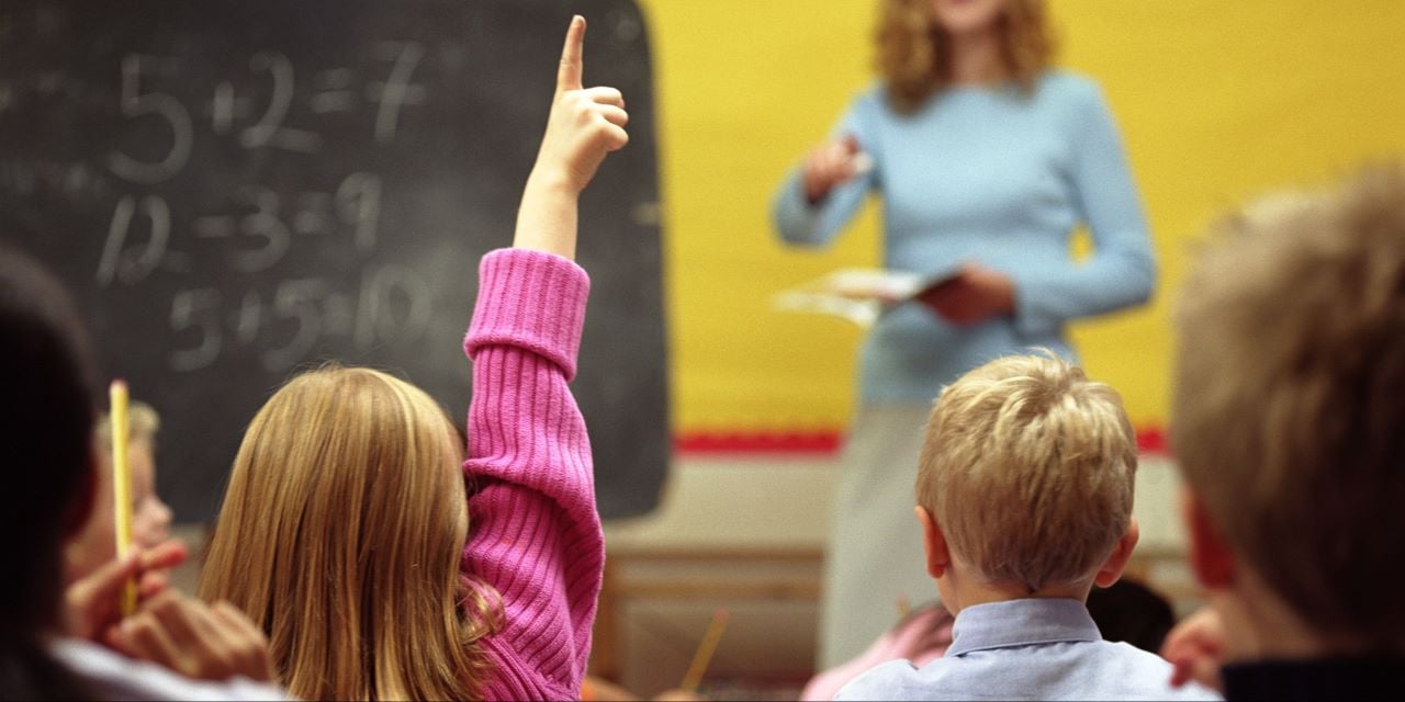 Student raining her hand in a classroom with a teacher at the front of the class.