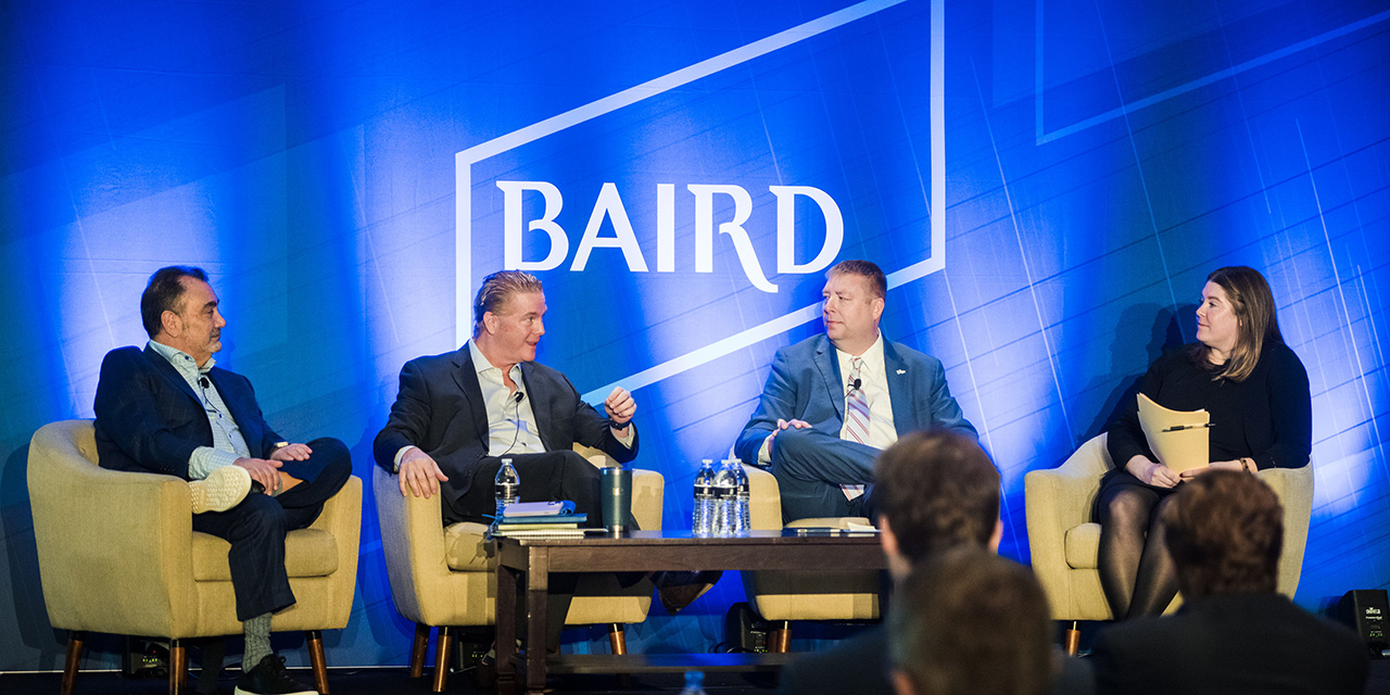 Three men and a woman on stage in front of a Baird logo talking during a panel presentation.