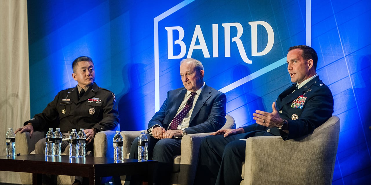 Three men sitting in chairs on stage during a panel discussion in front of a Baird logo.
