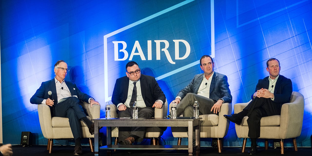 Four professionals on stage in front of a Baird logo during a presentation. 