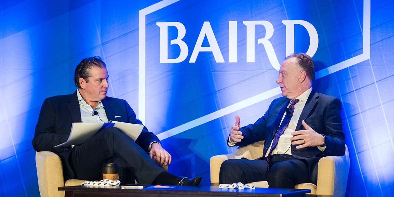 Two men sitting on stage talking in front of a Baird logo.