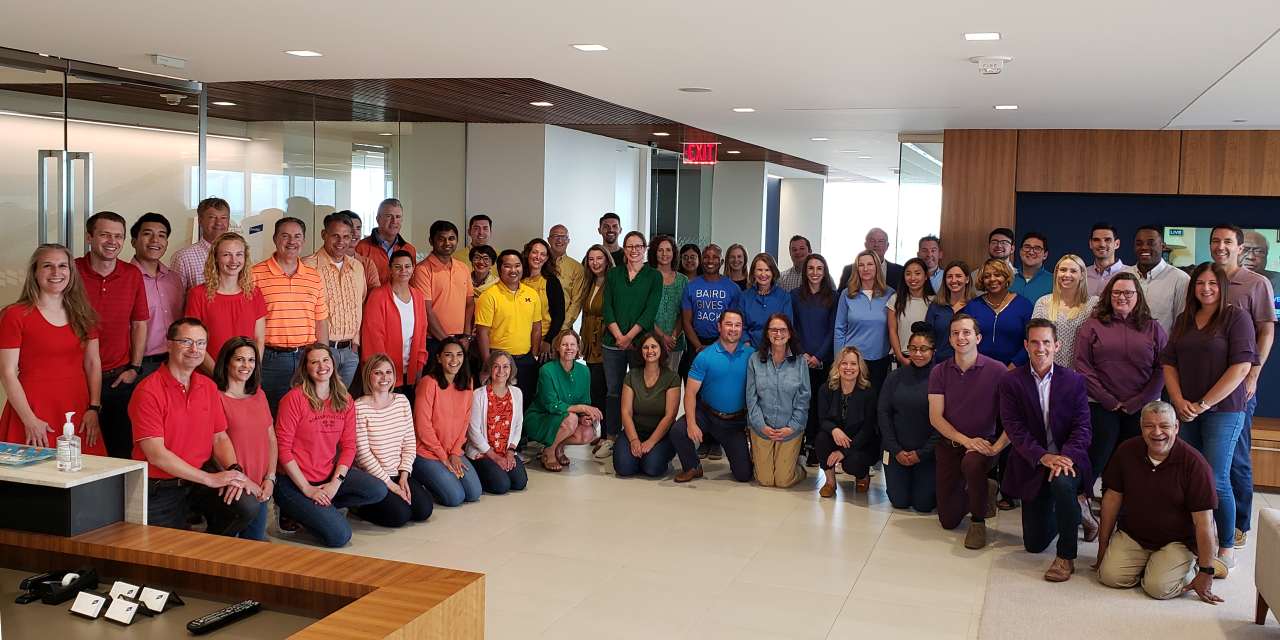 A large group of Baird associates wearing various, solid colored shirts stand in a rainbow arrangement